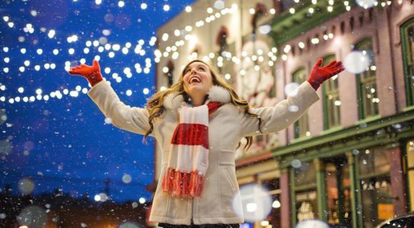 femme en manteau blanc avec écharpes blanche et rouge et gant blanc lève les bras sous guirlande lumineuse